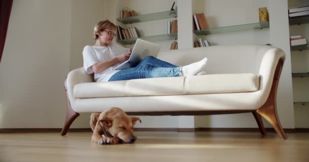 Kobieta patrzeć na ekranie laptopa siedzieć na kanapie, brązowy pies leży przy kanapie. Szeroki widok — Wideo stockowe