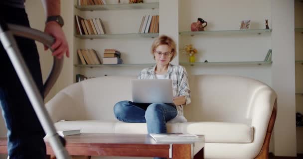 Moe vrouw zit op de bank, geïrriteerd terwijl een luidruchtige kleine dochter schreeuwt, springt op de bank en man doet de schoonmaak in de kamer. Slow motion shot — Stockvideo