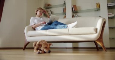 Kadın dizüstü bilgisayar ekranına bak kanepeye otur, kahverengi bir köpek kanepenin yanında yatıyor. Geniş çekim görüntüleri