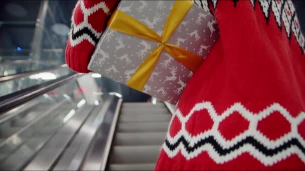 Ένας άντρας με ένα κόκκινο χριστουγεννιάτικο πουλόβερ κρατάει ένα δώρο στα χέρια του και ανεβαίνει στις κυλιόμενες σκάλες στο εμπορικό κέντρο. — Αρχείο Βίντεο