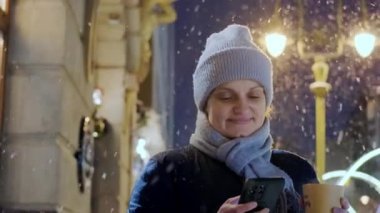 Mutlu, güler yüzlü bir kadın, Noel şehrinde, kar yağarken ve telefonla kahve içerken.