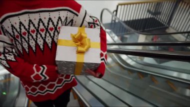 Kırmızı Noel kazaklı bir adam elinde bir hediye tutuyor ve alışveriş merkezinde yürüyen merdivene biniyor. Kapat.