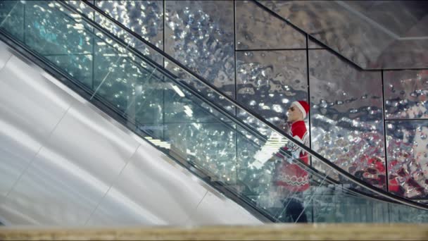 Un hombre con un suéter rojo de Navidad tiene un regalo en sus manos y monta en la escalera mecánica en el centro comercial. Tiro lateral — Vídeo de stock