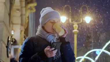 Mutlu, gülümseyen bir kadın, gece vakti kar yağarken ve telefon kullanırken eve servis kahvesini yudumluyor. Yavaş çekim.