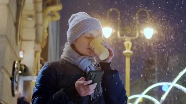 Mutlu, gülümseyen bir kadın, gece vakti kar yağarken ve telefon kullanırken eve servis kahvesini yudumluyor. Yavaş çekim. — Stok video