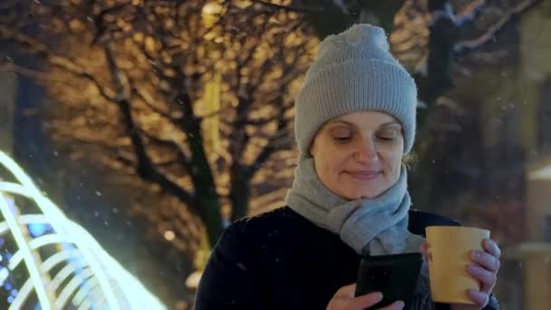 Щаслива усміхнена жінка п'є каву вночі в Різдвяному місті над снігом падає і використовує телефон Пересунути камеру — стокове відео