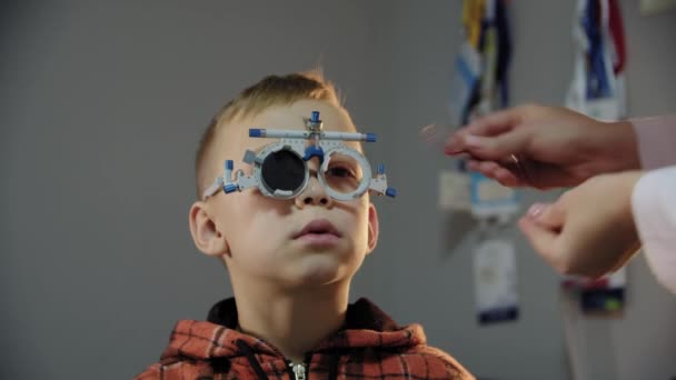 Van dichtbij gezien. De oogarts onderzoekt de patiënt tiener jongen oog en zet op een apparaat voor het selecteren van lenzen voor bril — Stockvideo