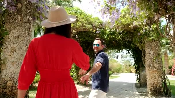 Mladý pár muž a žena v červených šatech prochází zahradou a s úsměvem se otočí. Zpomal. Široký pohled — Stock video