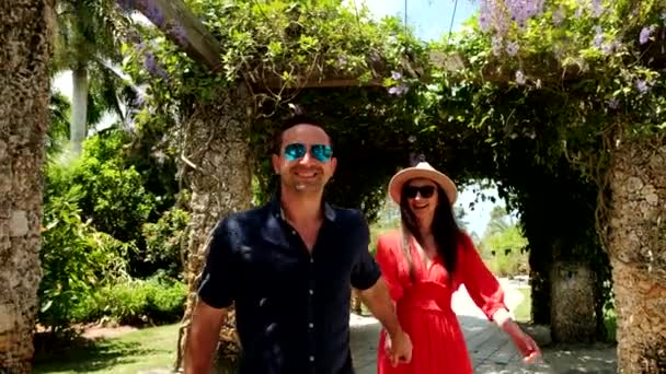 Unga par man och kvinna i röd klänning går genom trädgården och vänder sig om och ler. Sakta i backarna. Bredbildsfilm — Stockvideo