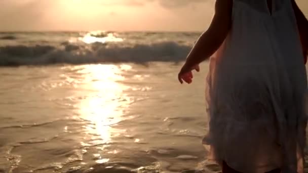 Una niña con un vestido blanco está parada a lo largo de la playa. De cerca. — Vídeos de Stock
