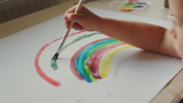 La niña sostiene un pincel en la mano y pinta un arco iris con pinturas de colores sobre papel blanco. Movimiento lento — Vídeo de stock