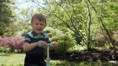 Küçük çocuk güneşli bir günde parkta scooter sürüyor. Yavaş çekim