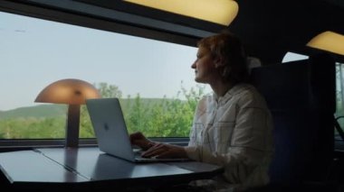 Kadın, güneşli bir günde trende pencerenin yanında otururken dizüstü bilgisayarla çalışıyor. Geniş açı