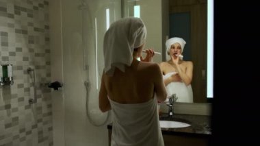 Kafasında beyaz banyo havlusu olan kadın aynaya bakarak dişlerini fırçalıyor. Yüksek kalite 4k görüntü