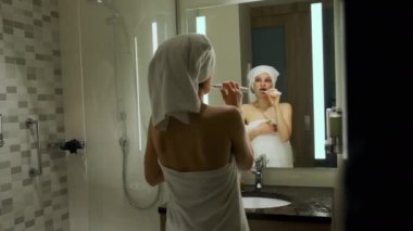 Kafasında beyaz banyo havlusu olan kadın aynaya bakarak dişlerini fırçalıyor. Yüksek kalite 4k görüntü