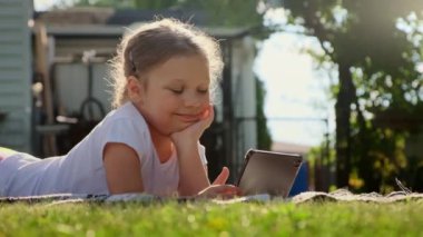 Kaygısız kız tablette oynuyor. Evin yakınındaki çimlerin üzerinde yatıyor, güneş saçlarını aydınlatıyor. Yüksek kalite 4k görüntü