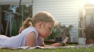 Kaygısız kız tablette oynuyor. Evin yakınındaki çimlerin üzerinde yatıyor, güneş saçlarını aydınlatıyor. Yüksek kalite 4k görüntü