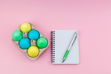 Modaya uygun koyu pembe arka planda boş not defteri ve kalem. Fikir mesajı, liste ve ilham için not defteri. Not defterinin yanında çok renkli Paskalya yumurtaları olan tepsi. Tasarımın için model al..