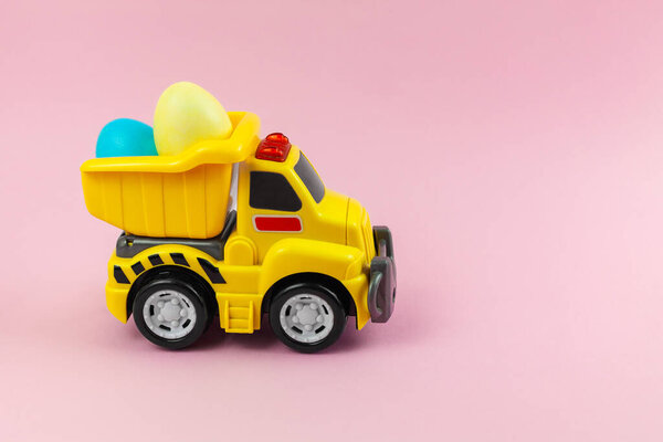 A toy yellow truck carrying Easter eggs in pastel colors. Easter card with place for text on a pink background. Easter food delivery