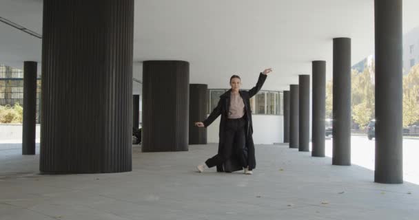 Attractive businesswoman in formal black trench is dancing outside the office, financial district, modern constructivist building with columns on background. The concept of urban city scene. — ストック動画