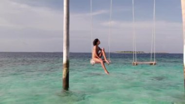 Maldivler adasının ve salıncaktaki genç kadının havadan çekilmiş görüntüleri. Okyanus, orman ve ufuk çizgisinin panoramik görüntüsü. Turizm ve tatil kavramı