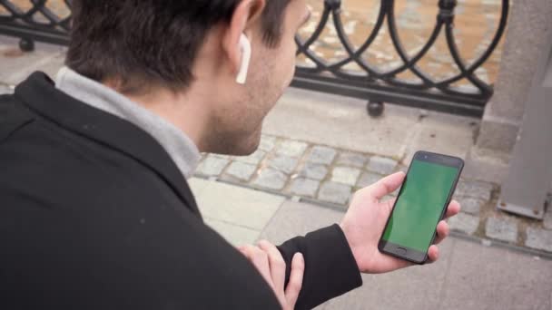 商人使用带有绿色屏幕 彩色键和无线耳机的智能手机 坐在附近的河里 肩膀上的一枪 — 图库视频影像