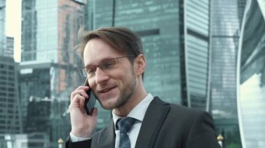 Yakışıklı iş adamı yönetici patron iş ortağıyla telefonda konuşuyor. Arka planda şirket konsepti gökdelenleri.