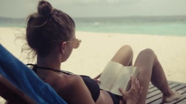 Mayolu çekici bir kız elinde kitapla kumsalda güneşlenir. Önümüzde sakin mavi bir deniz var..