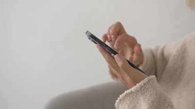 Yan görüş. El ele tutuşan ve evdeki beyaz odada akıllı telefon kullanan bir kadın internete mesaj yazıyor. Kapat..