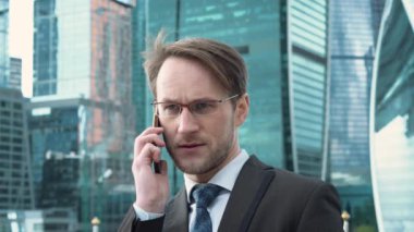Yakışıklı iş adamı yönetici patron iş ortağıyla telefonda konuşuyor. Arka planda şirket konsepti gökdelenleri.