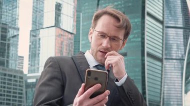 Yakışıklı iş adamı, video konferans görüşmesiyle cep telefonuyla konuşuyor. Arka planda şirket konsepti gökdelenleri.