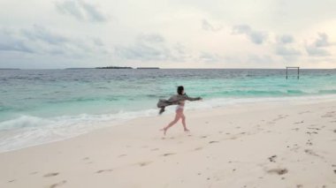 Neşeli kadının Maldivler 'deki kumsalda, gökyüzünde ve turkuaz Hint okyanusu dalgalarında hızla koşarken çekilmiş görüntüsü. Turizm ve mutluluk kavramı