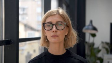 Ofis odasının arka planında gergin duygular olan gözlüklü sarışın kadın portresi. Tarafsız bir ifadeyle kameraya bakan çekici bir stil sahibi. İş kadını kavramı. 