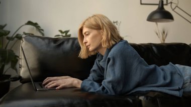 Genç bir kadın dizüstü bilgisayar kullanıyor, oturma odasında kanepede dinleniyor. Kot pantolonlu bir kadın konsantre olmuş, bir cihaza mesaj atıyor. Evden çalışma ve boş zaman kavramı. 