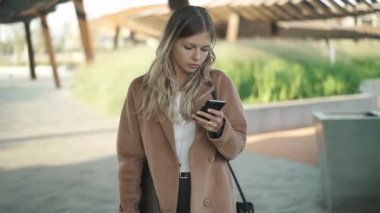 Bir palto giyiyor sarışın saçlı Genç ve güzel kadın sonbahar sokakta duran ve onun smartphone vasıl arıyorum. Sosyal medya ve sokak moda kavramı. Ağır çekim portre çekim izleme.