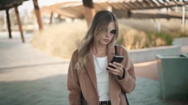 Sarı saçlı bir ceket ve gözlük çekici genç kadın sonbahar street yürüyüş ve onun smartphone vasıl arıyorum. Ağır çekim portre çekim izleme.
