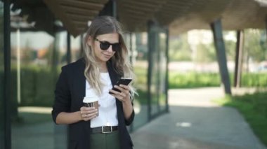 Gidip onu akıllı telefon ekranına bakarak gülen çekici genç kadın güneş gözlüğü ve kahve tutan takım elbise uzun sarı saçlı. Sosyal medya Slider ağır çekim portre çekim kavramı.