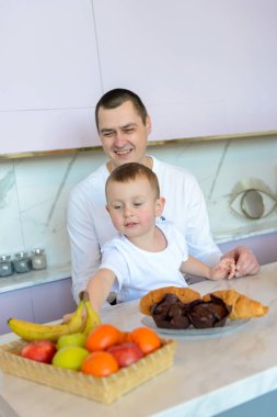 Mutlu baba ve oğul, modern mutfaktaki masada meyvelerle kahvaltı yapıyorlar.