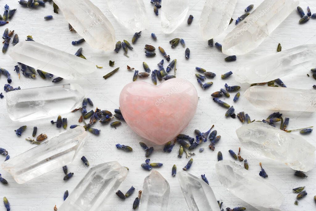 A close up image of several clear quartz crystals with a heart shaped rose quartz crystal and dried lavender flowers. 