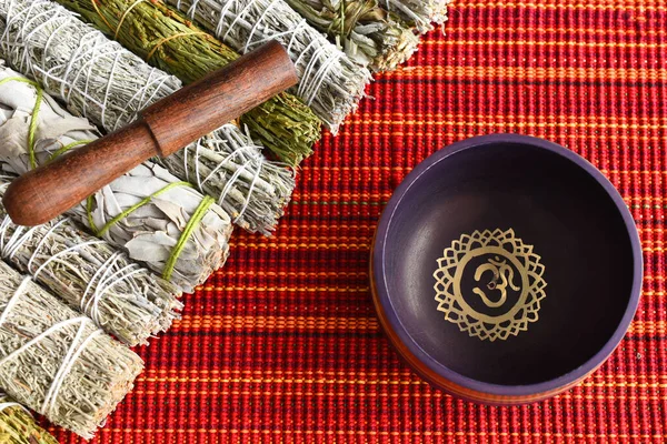 A top view image of a purple meditation singing bowl with a variety of healing smudge sticks on a red and orange background.