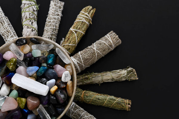 A top view image of a bowl of healing crystals surrounded by various smudge sticks on a black background. 