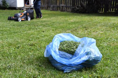 A close up image of a large blue plastic bag used for recycling grass cuttings with a lawn mower in the background. 