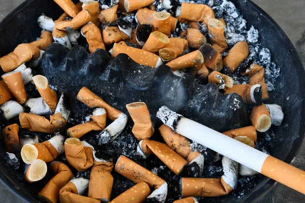 A close up image of a dirty unhealthy ashtray with a burning cigarette.