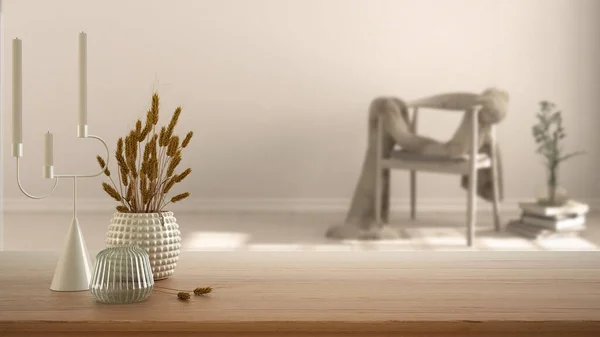 Wooden table, desk or shelf close up with ceramic and glass vases with dry plants, straws over blurred view of scandinavian living room with armchair, modern interior design concept
