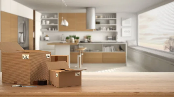 Wooden table, desk or shelf with stack of cardboard boxes over blurred view of minimalist kitchen with island, modern interior design, moving house concept with copy space