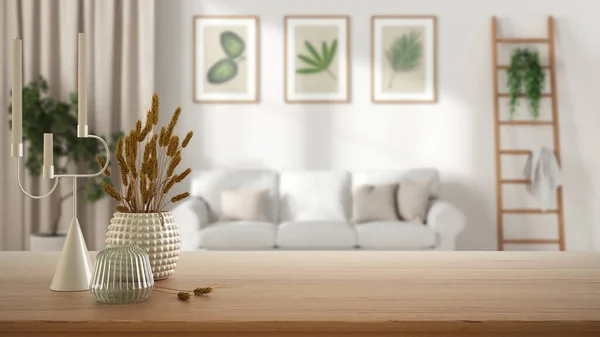 Wooden table, desk or shelf close up with ceramic and glass vases with dry plants, straws over blurred view of scandinavian living room with sofa, modern interior design concept
