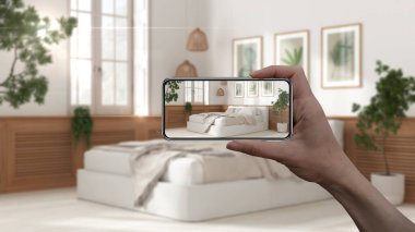El ele tutuşan akıllı telefon, AR uygulaması, gerçek evde mobilya ve iç tasarım ürünleri simülasyonu, mimar tasarım konsepti, bulanık arka plan, yatak ve dekorlu beyaz yatak odası
