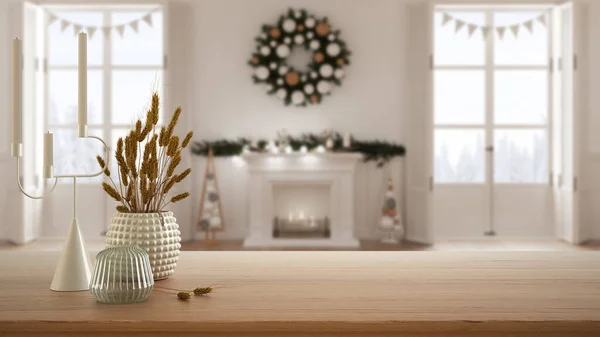 Wooden table, desk or shelf close up with ceramic and glass vases with dry plants, straws over blurred view of Christmas living room with fireplace, modern interior design concept
