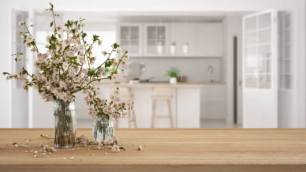Wooden table, desk or shelf close up with branches of cherry blossoms in glass vase over blurred view of scandinavian white kitchen, boho interior design concept