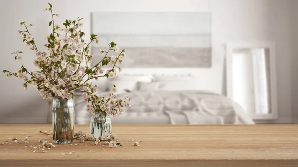 Wooden table, desk or shelf close up with branches of cherry blossoms in glass vase over blurred view of scandinavian white bedroom, boho interior design concept